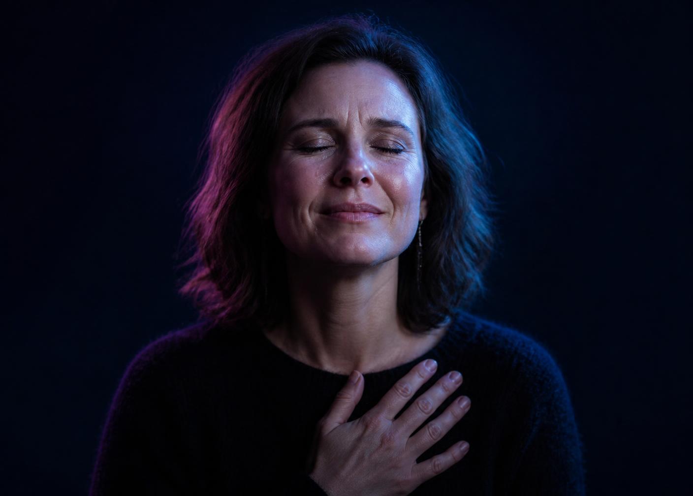 A woman with her eyes closed and a hand on her heart, lit in violet and cyan light